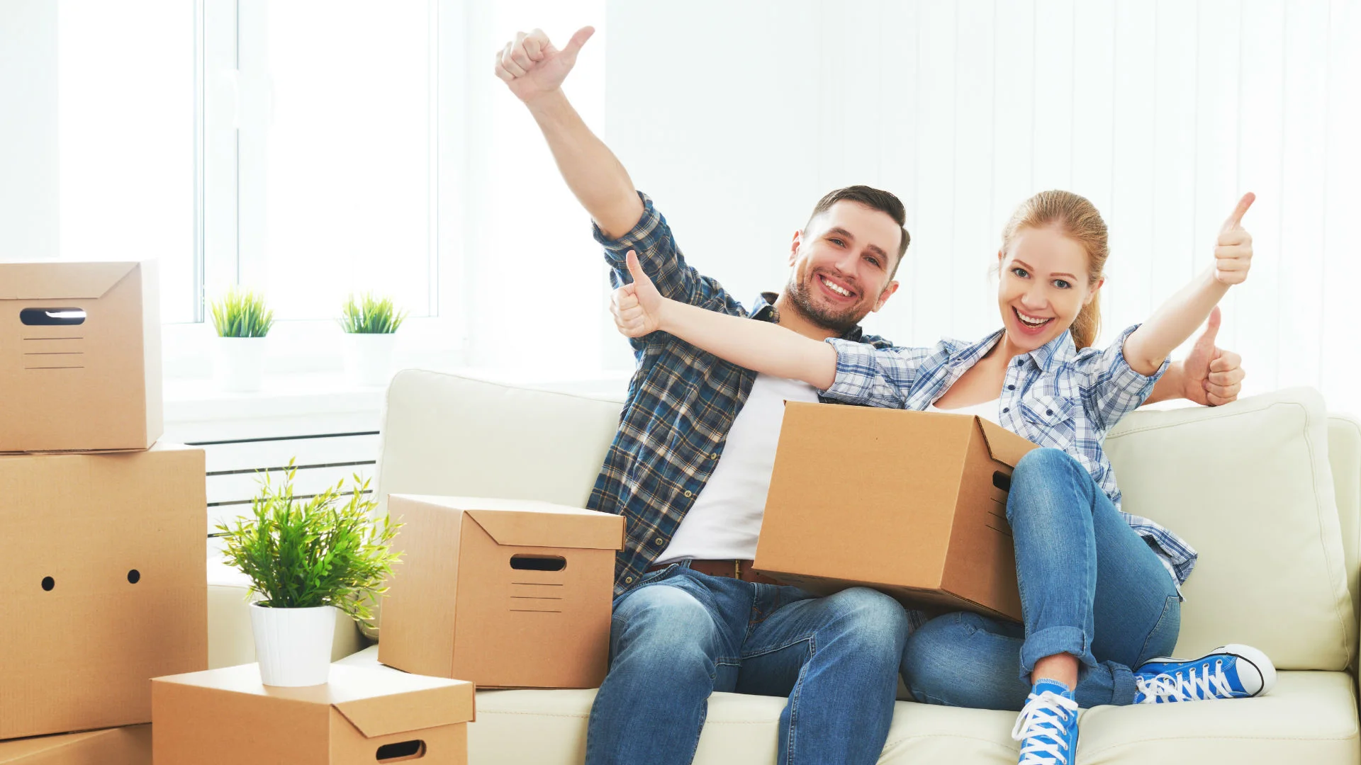 Happy couple giving thumbs up after packing for their move, surrounded by cardboard boxes.